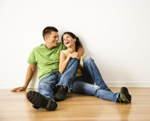 Attractive young adult couple sitting close on hardwood floor in home smiling and laughing.